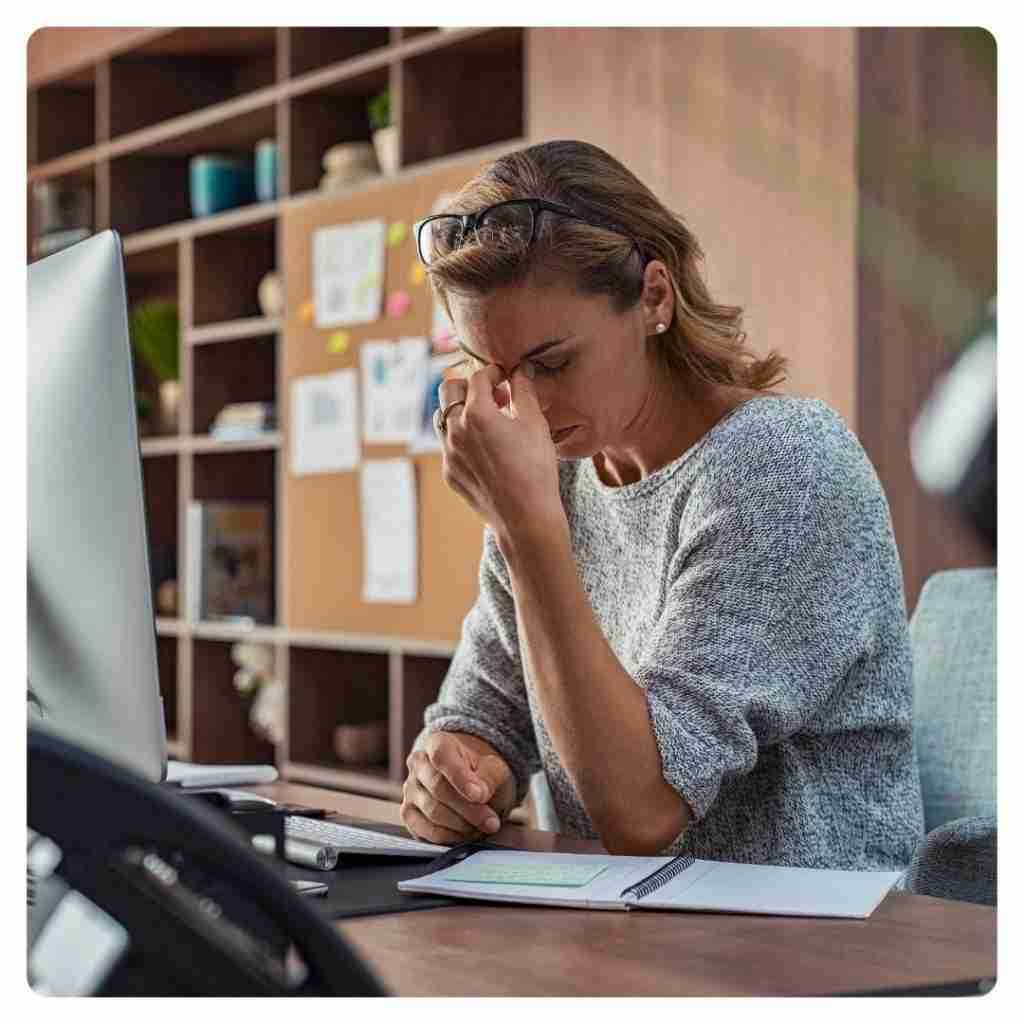 Spine Trouble from Sitting - female at her desk at work pinching her forehead between the eyes indicating a headache