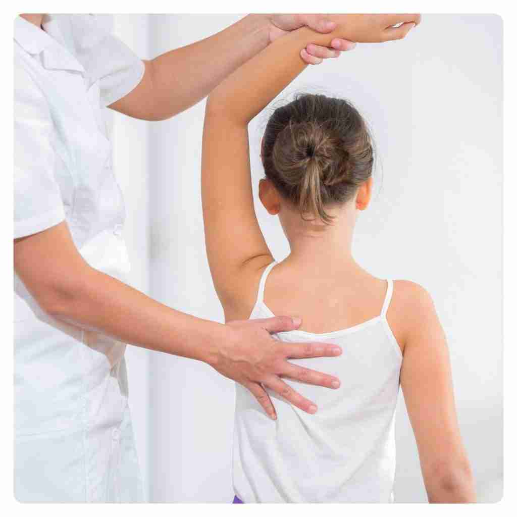 Benefits of Chiropractic Adjustments - chiropractor assessing the back of a young female patient by placing hand on back and raising her left arm to feel for misalignments
